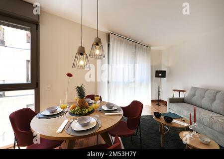 Wohnzimmer einer Ferienwohnung mit einem runden Holztisch mit einem Willkommensfrühstück mit Säften und Obst Stockfoto