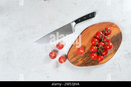 Leuchtende kleine rote Tomaten mit grünen Reben auf Holzhacke, großes Küchenmesser in der Nähe, weißer Steintisch unter dem Tisch, Blick von oben Stockfoto