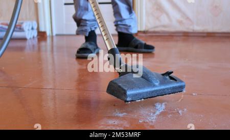 Staubsaugerrohr mit schwarzer Kunststoffbürste saugt grauen Staub von einem braunen Holzboden und Männerfüßen auf einem verschwommenen Hintergrund auf Stockfoto