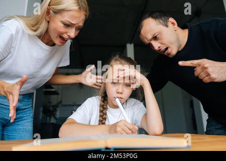 Porträt von wütenden jungen Eltern, die zusammen schimpfen und schimpfen, faule kleine Tochter, die am Tisch sitzt, Hausaufgaben macht, traurig in die Kamera schaut. Stockfoto