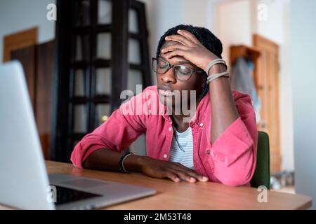 Unterdrückter trauriger afroamerikanischer Mann mit geschlossenen Augen sitzt am Tisch mit Laptop und fühlt sich ausgebrannt Stockfoto