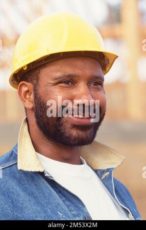 Ein Afroamerikaner mit Bart und Schutzhelm auf einer Baustelle, der in die Ferne blickt Stockfoto