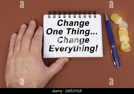 Geschäftskonzept. Auf einer braunen Oberfläche befinden sich Münzen, ein Stift und ein Notizblock mit der Aufschrift „eine Sache ändern und alles ändern“. Das Notizbuch heißt "Hol" Stockfoto