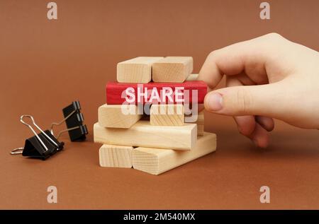 Geschäftskonzept. Klemmen und Holzblöcke liegen auf einer braunen Oberfläche, eine Person nimmt einen roten Block mit der Aufschrift „Teilen“ heraus Stockfoto