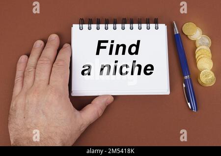 Geschäftskonzept. Auf einer braunen Oberfläche befinden sich Münzen, ein Stift und ein Notizblock mit der Aufschrift – Finden Sie eine Nische. Das Notizbuch hält eine Hand. Stockfoto