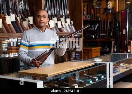 Gutaussehender, positiver erwachsener Mann im Jagdgeschäft mit seltenem Gewehr in den Händen Stockfoto