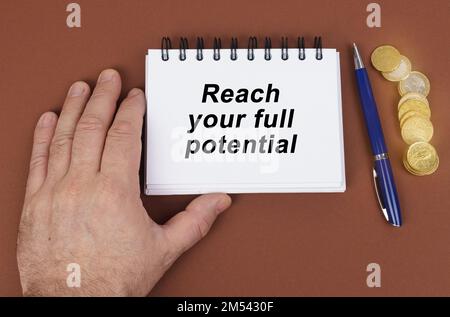 Geschäftskonzept. Auf einer braunen Oberfläche befinden sich Münzen, ein Stift und ein Notizblock mit der Inschrift – so erreichen Sie Ihr volles Potenzial. Das Notizbuch hält eine Hand. Stockfoto