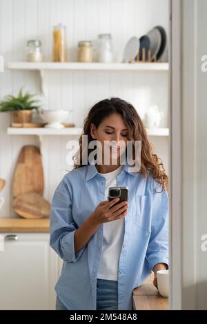 Junge, angenehme lächelnde Frau, die in der Küche stand, ihr Smartphone hielt und sich ein Kochvideo anschaute Stockfoto