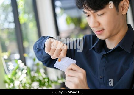 Gestresster und wütender junger asiatischer Mann, der seine Kreditkarte zerbricht, seine Kreditkarte zerstört, finanzielle Probleme hat. Stockfoto