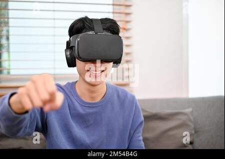Fröhlicher und gutaussehender junger asiatischer Mann mit VR-Brille, der in seinem Wohnzimmer VR-Spiele spielt. Stockfoto