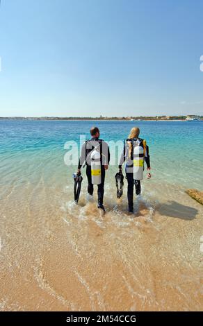 Junges Paar, junger Mann und junge Frau, die zusammen mit einer Tauchausrüstung für das Tauchen Seite an Seite über den Strand im Roten Meer, Makadi, spazieren gehen Stockfoto