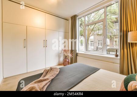 Ein Schlafzimmer mit einem Bett und Kleiderschränken vor einem großen Fenster mit Vorhängen an der Außenwand Stockfoto