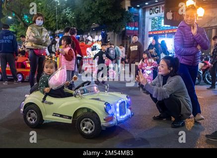 Eine Mutter macht ein Foto ihrer kleinen Tochter, die in einem Spielzeugauto am Wochenende auf der Straße um den Hoan Kiem See im Zentrum von Hanoi, Vietnam, herumläuft. Stockfoto