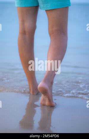 Eine vertikale Nahaufnahme der Füße einer Frau, die auf einem Sandstrand gegen Meereswellen läuft Stockfoto