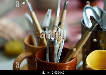 Verschiedene Werkzeuge zum Schnitzen von Töpferwaren in Tassen, die in der Werkstatt auf einem Holztisch mit professionellen Instrumenten aufgestellt werden. Stockfoto