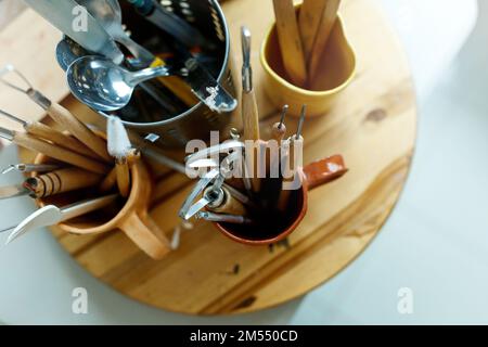 Verschiedene Werkzeuge zum Schnitzen von Töpferwaren in Becher auf Holztisch in der Werkstatt mit professionellen Instrumenten, Draufsicht. Stockfoto