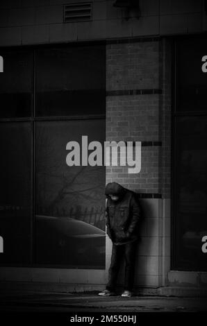 Eine vertikale Graustufenaufnahme einer einsamen, depressiven Person, die unter einer Mauer auf der Straße steht Stockfoto