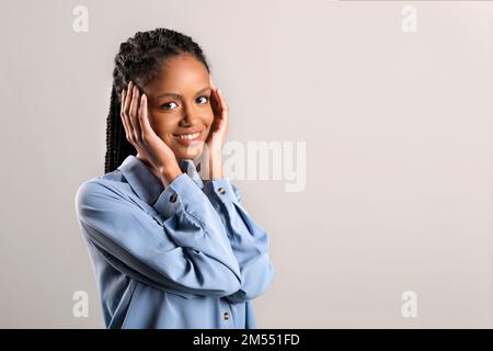 Glückliche junge schwarze Frau in blauem Hemd mit langen Afro-Geflechten, die mit einem Lächeln in die Kamera schaut und sich mit dem Gesicht vor grauem Hintergrund berührt Stockfoto