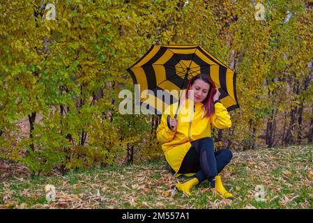 Ein Mädchen sitzt auf dem Rasen in einem gelben Regenmantel und einem gestreiften Regenschirm Stockfoto