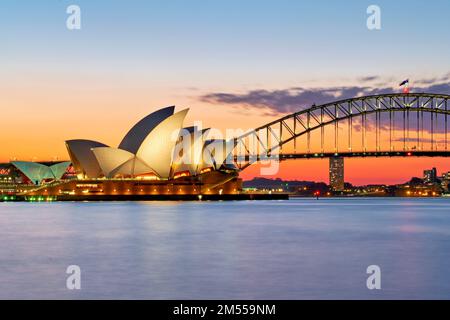 Australien. Das Opernhaus von Sydney und die Harbour Bridge, die bei Sonnenuntergang beleuchtet wird, bieten einen lebendigen Sonnenuntergang über dem Wasser Stockfoto