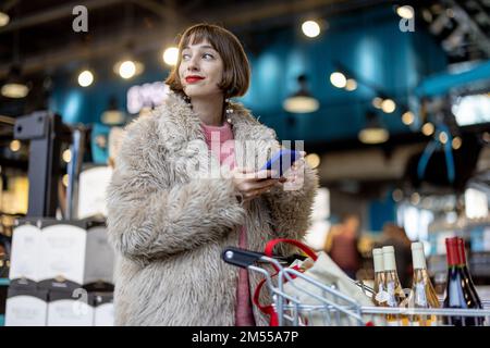 Frau mit Telefon und Wein im Supermarkt Stockfoto