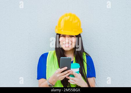 Porträt einer lateinischen Ingenieurin mit Schutzhelm und reflektierender Weste, die mit einer Wand im Hintergrund auf der Straße steht, lächelt, sie hält Stockfoto