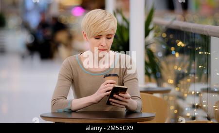 Erwachsene Frau mittleren Alters mit kurzer Frisur, sitzt am Café-Tisch und trinkt Kaffee, Tee, schaut sich die Handy-App an und chattet mit ihr Stockfoto