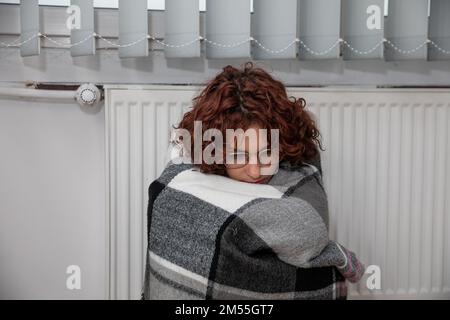 Ein Mädchen, eingewickelt in einen dicken Schal, sitzt neben einem warmen Heizkörper. Zentralheizung. Stockfoto