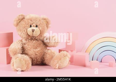 Kinderspielzeug-Hintergrund. Spielzeugteddybär, pastellfarbener Holzregenbogen und pinkfarbene Ziegelwürfel auf pastellrosa Hintergrund. Vorderansicht Stockfoto