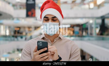 Indischer ethnischer Mann männlich in medizinische Maske in Weihnachten Weihnachten Weihnachtsmann hat Wetten mit Handy Casino Wette Online Bank Zahlung Kreditservice schnell Stockfoto