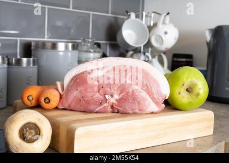 Rohes, nicht gekochtes Schweinebein-Schultergelenk mit gekochtem Apfelknips und Karotten auf einem Holzbrett Stockfoto