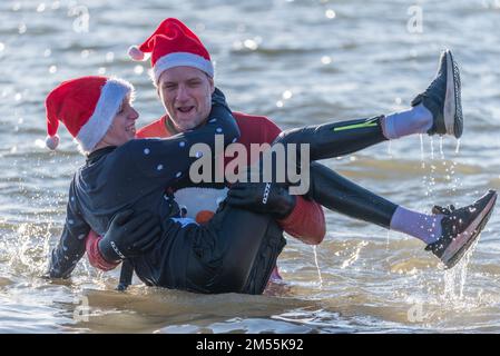 Jubilee Beach, Marine Parade, Southend on Sea, Essex, Großbritannien. 26. Dezember 2022. Wie es Tradition an Orten am Meer geworden ist, fand ein „Boxing Day Dip“ in der kalten, rauen Themsenmündung in Southend on Sea in der Nähe des Stadtpiers statt und sammelte Gelder für die örtliche Royal National Lifeboat Institution. Etwa 400 Personen gingen zum Wasser, das etwa 6 Grad Celsius betrug. Viele der mutigen Schwimmer trugen festliche Kleidung. Ein Mann, der einen Jungen trägt Stockfoto