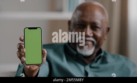Ein alter afroamerikanischer birassischer Mann zu Hause, der auf der Couch sitzt, ein Geschäftsmann im Büro, der ein Smartphone mit leerem grünen Bildschirm zeigt Stockfoto