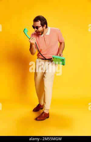 Porträt eines stilvollen Mannes mit Schnurrbart, Geschäftsmann in Vintage-Kleidung, der vor einem Retro-Telefon schreit, isoliert auf gelbem Hintergrund. Retro-Style Stockfoto