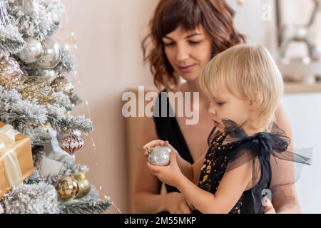 Eine Mutter mit einer 2-jährigen Tochter schmückt den Weihnachtsbaum. Beide tragen schwarze Kleider, die Tochter hängt einen Ball an den Weihnachtsbaum. Stockfoto