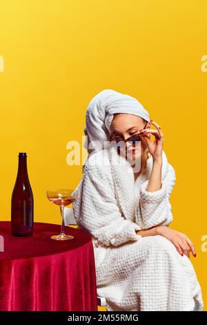 Junge Frau im Bademantel, sitzt und trinkt Champagner auf gelbem Hintergrund. Feier Stockfoto