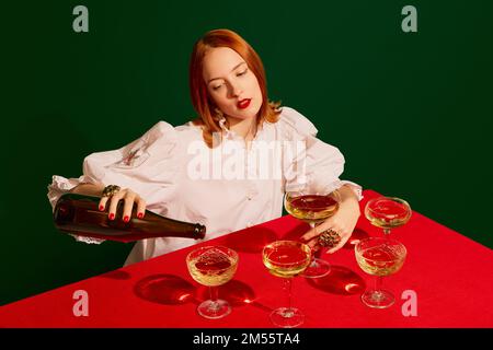 Junge Rothaarige in weißer Kleidung, die am Tisch sitzt und Champagner in Glas mit grünem Hintergrund gießt. Stockfoto