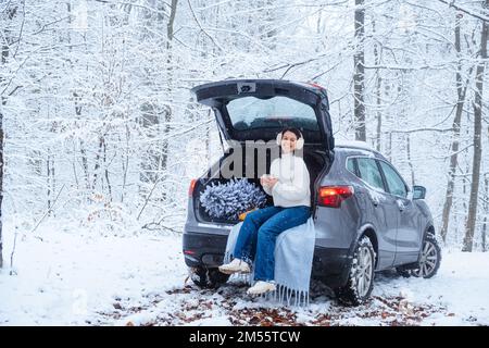 Eine junge Frau sitzt in einem Kofferraum mit einem Weihnachtsbaum im Wald Stockfoto