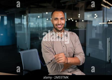 Porträt eines afroamerikanischen Geschäftsmanns im Büro, Mann im Hemd, der am Fenster lächelt und in die Kamera schaut, Programmierer, der in einem Entwicklungsunternehmen arbeitet. Stockfoto
