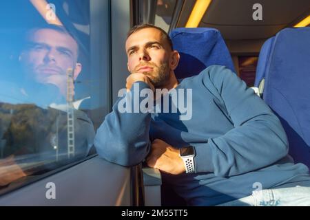 Der junge Mann hält seine Hand am Kinn und blickt aus dem Fahrgastraum aus dem Zugfenster und genießt den Blick auf die von der Sonne beleuchtete Natur Stockfoto