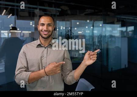 Hispanischer Geschäftsmann im Büro, der lächelt und in die Kamera schaut, während er steht und mit den Händen zur Seite zeigt, ein Mann in einem lässigen Hemd nahe dem Fenster. Stockfoto