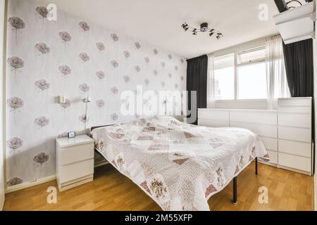 Ein Schlafzimmer mit Blumentapeten an den Wänden und Holzfußboden vor dem Bett, es gibt ein Fenster zum Stockfoto