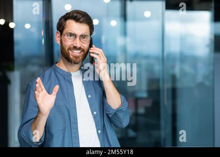 Porträt eines erwachsenen, erfolgreichen Geschäftsmannes im Büro, der Mann lächelt und in die Kamera am Fenster schaut, während er steht und fröhlich spricht und am Telefon lächelt. Stockfoto