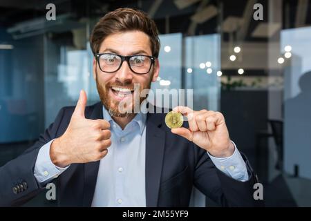 Porträt eines reifen erfolgreichen Geschäftsmanns Investor, Mann zeigt kryptowährungsfähige Goldmünzen vor der Kamera und Daumen hoch, Mann arbeitet an der Börse im Büro online verwendet Laptop. Stockfoto