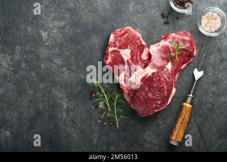 Rohes Rindersteak. Marmoriertes rohes Ribeye-Steak in Herzform mit Rosmarin, Salz und Pfeffer auf dem Schneidebrett auf dunklem Betonhintergrund. Rohes Rindfleisch St. Stockfoto