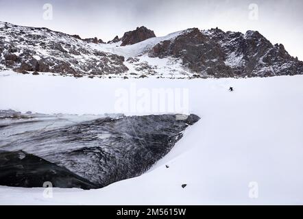 Skifahrer mit rotem Rucksack im Helm und Maske in den hohen schneebedeckten Bergen in der Nähe der Eismauer mit Schneespritzern. Extremer Outdoor-Sport im Winter Stockfoto