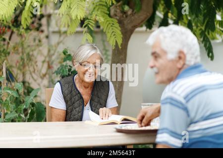 Liebevolle ältere Ehepartner, die sich im Garten ausruhen und Tage genießen, draußen sitzen. Frau liest Buch, Mann vor ihr. Stockfoto