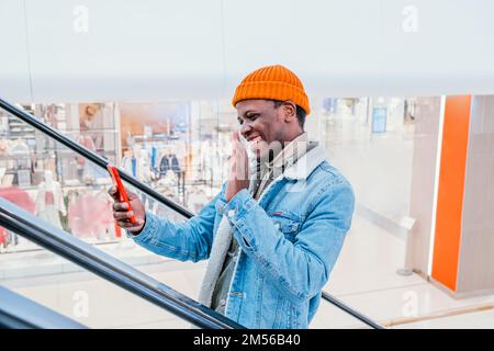 Fröhlicher junger afroamerikanischer Mann in stilvoller Kleidung benutzt Video-Chat auf dem Mobiltelefon, der die Rolltreppe in der Seitenansicht des Einkaufszentrums hochfährt Stockfoto