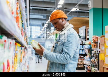 Nachdenklicher afroamerikanischer Mann wählt Saft in einer Papierverpackung, die in der Nähe einer großen Ablage in der Seitenansicht des modernen Supermarktbereichs steht Stockfoto