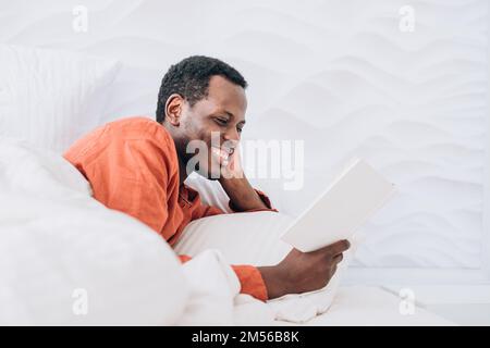 Ein positiver afroamerikanischer Mann im Schlafanzug liest interessante Romane, die in einem bequemen Bett unter einer weichen Decke in der Nähe des Zimmers liegen Stockfoto